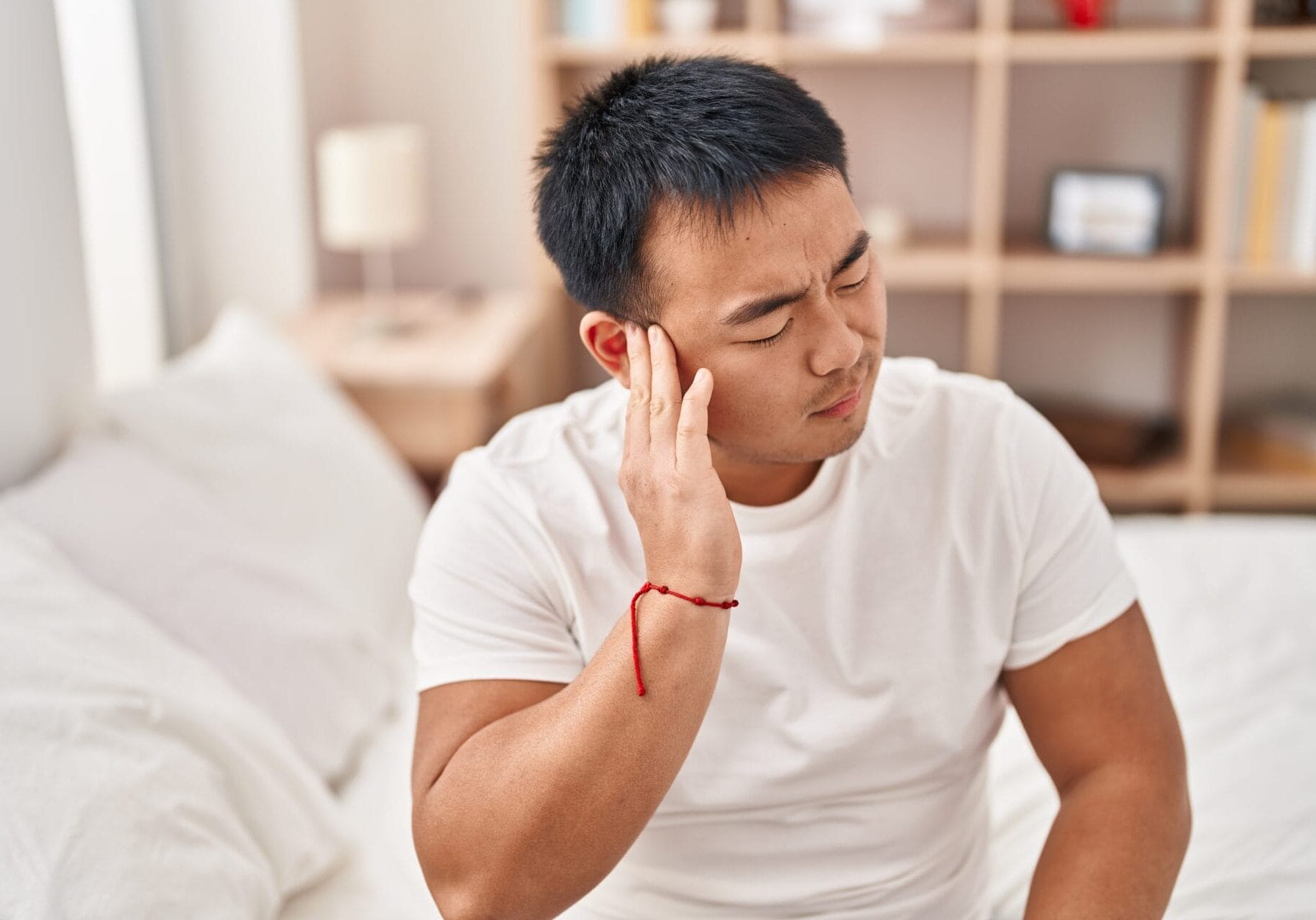 Young chinese man suffering for ear pain sitting on bed at bedroom Young chinese man suffering for ear pain sitting on bed at bedroom